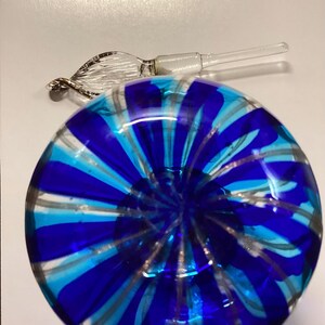 May include: A round glass perfume bottle featuring a vibrant pattern of alternating royal blue and turquoise stripes radiating from the centre. A clear glass stopper with a twisted, leaf-like decorative handle rests beside the bottle.