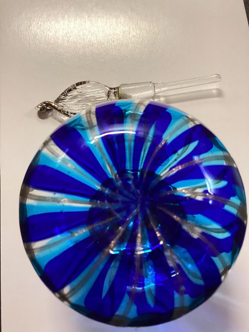 May include: A round glass perfume bottle featuring a vibrant pattern of alternating royal blue and turquoise stripes radiating from the centre. A clear glass stopper with a twisted, leaf-like decorative handle rests beside the bottle.