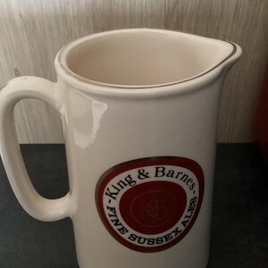 Op de afbeelding: Cr&egrave;mekleurige keramische kan met een handvat en een schenktuit. De kan heeft een cirkelvormig embleem met de tekst "King & Barnes FINE SUSSEX ALES" in een rood en goud ontwerp. De rand heeft een gouden rand.