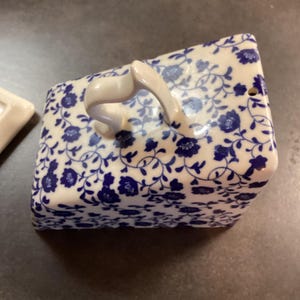 May include: A rectangular ceramic butter dish cover featuring a white base with a dense blue floral pattern. The lid has a curved, arched handle on top and a small circular vent hole on one side, showcasing a glossy finish and intricate vine details.