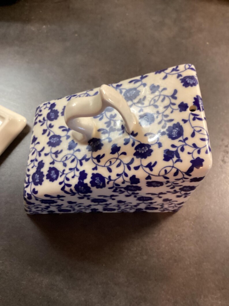 May include: A rectangular ceramic butter dish cover featuring a white base with a dense blue floral pattern. The lid has a curved, arched handle on top and a small circular vent hole on one side, showcasing a glossy finish and intricate vine details.
