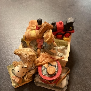 May include: A detailed figurine featuring a teddy bear, a toy train, a drum, and toy soldiers. The train is red and black with the number 3 on the side. The teddy bear holds a teacup. The figurine is on a small base.
