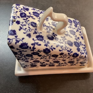 May include: A white ceramic butter dish with a blue floral pattern covering the lid. The lid features a curved handle on top and a small hole on one side. The base is a rectangular white tray with a raised edge to hold the lid securely in place.