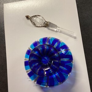 May include: A round glass paperweight featuring alternating stripes of deep blue, light blue, and clear glass, accompanied by a separate clear glass stopper with a textured, leaf-like finial and a smooth, tapered stem.