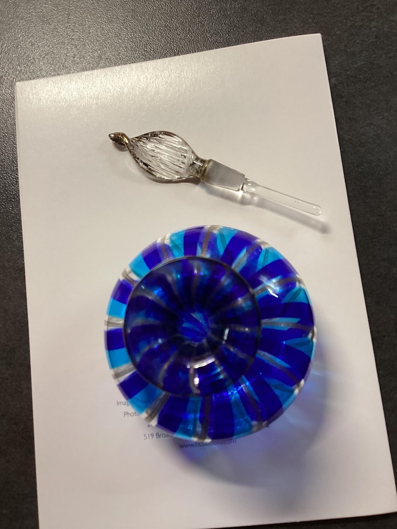 May include: A round glass paperweight featuring alternating stripes of deep blue, light blue, and clear glass, accompanied by a separate clear glass stopper with a textured, leaf-like finial and a smooth, tapered stem.