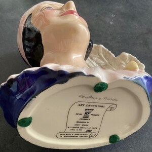 May include: A ceramic Art Deco girl figurine with a pink face, red lips, and black hair. The figurine has a blue and white collar and is marked "Feathers Handle" and "ART DECCO GIRL" with the text "A Limited Edition of 1,000 This is No 292".