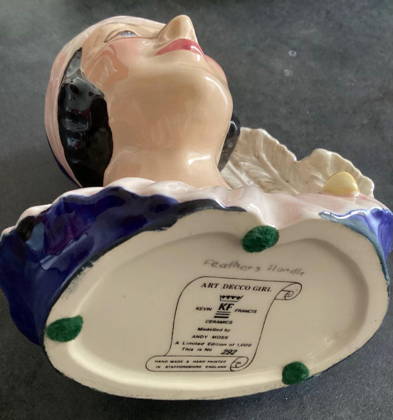 May include: A ceramic Art Deco girl figurine with a pink face, red lips, and black hair. The figurine has a blue and white collar and is marked "Feathers Handle" and "ART DECCO GIRL" with the text "A Limited Edition of 1,000 This is No 292".