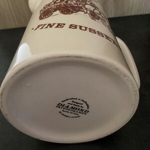 Op de afbeelding: Cr&egrave;mekleurige keramische kan met een bruine illustratie van een vintage voertuig en de woorden "FINE SUSSEX". De kan heeft een handvat en een brede basis. De onderkant van de kan heeft de tekst "DIAMOND PRINTCERAMICS".