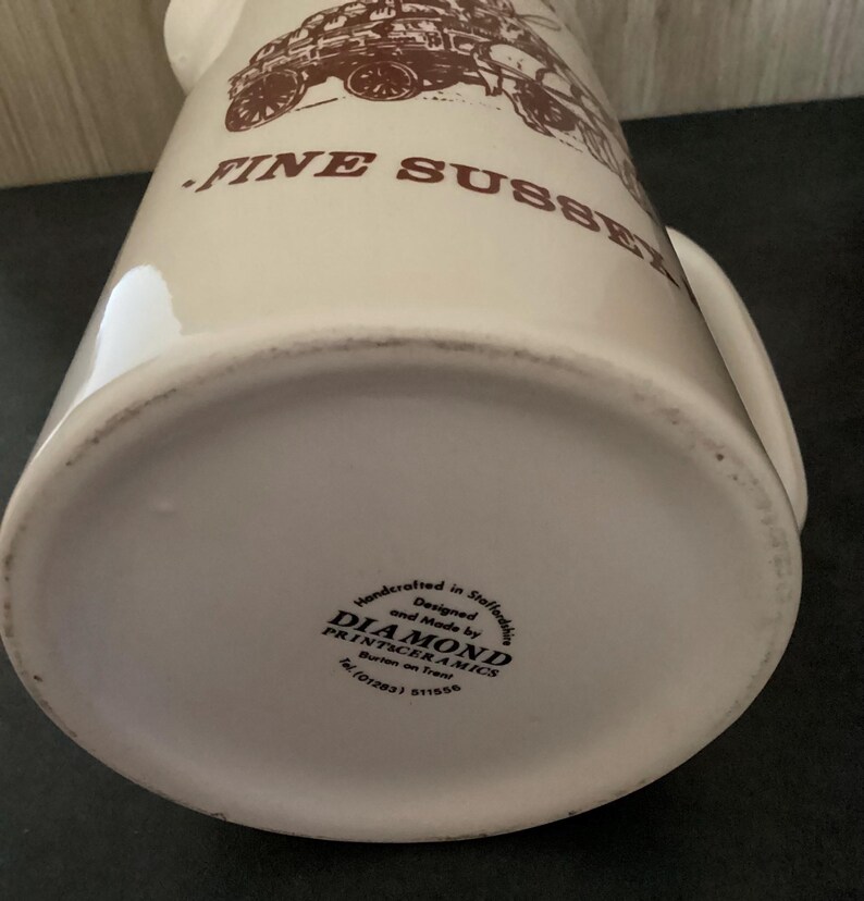 Op de afbeelding: Cr&egrave;mekleurige keramische kan met een bruine illustratie van een vintage voertuig en de woorden "FINE SUSSEX". De kan heeft een handvat en een brede basis. De onderkant van de kan heeft de tekst "DIAMOND PRINTCERAMICS".