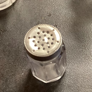 Könnte beinhalten: Ein klarer Salzstreuer aus Glas mit einem silbernen Metalldeckel mit mehreren kleinen Löchern. Der Streuer hat einen facettierten, sechseckigen Sockel und einen zylindrischen Körper. Der Deckel hat einen dekorativen, perlenbesetzten Rand.