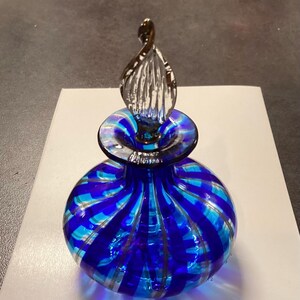 May include: A round glass perfume bottle featuring alternating vertical stripes of deep cobalt blue and translucent light blue. The bottle has a clear, twisted, flame-shaped glass stopper that sits securely in the narrow neck of the vessel.