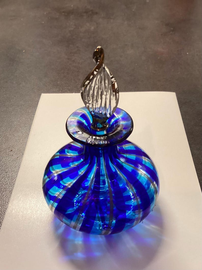May include: A round glass perfume bottle featuring alternating vertical stripes of deep cobalt blue and translucent light blue. The bottle has a clear, twisted, flame-shaped glass stopper that sits securely in the narrow neck of the vessel.