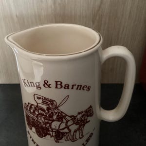 Op de afbeelding: Cr&egrave;mekleurige keramische kan met een handvat en een schenktuit. De kan heeft een bruine illustratie van een paardenkar en de tekst "King & Barnes Fine Sussex Ales".