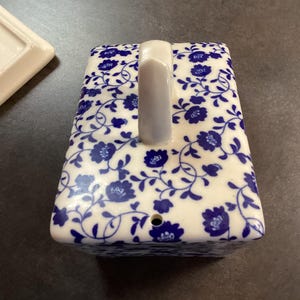May include: A rectangular ceramic butter dish lid featuring a white base with a blue floral pattern. The lid has a central vertical handle and a small circular hole near the bottom edge. A separate white ceramic base piece is partially visible.