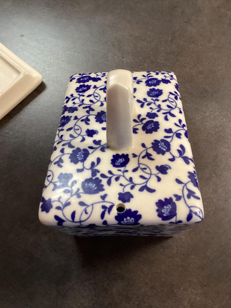 May include: A rectangular ceramic butter dish lid featuring a white base with a blue floral pattern. The lid has a central vertical handle and a small circular hole near the bottom edge. A separate white ceramic base piece is partially visible.