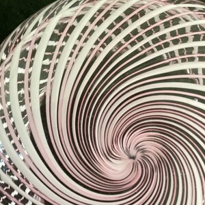 May include: A clear glass object featuring a swirling pattern of opaque white and soft pink lines that radiate from a central point, creating a dynamic vortex effect across the curved surface of the transparent material.