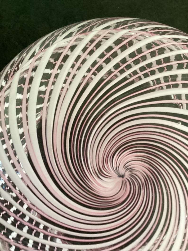 May include: A clear glass object featuring a swirling pattern of opaque white and soft pink lines that radiate from a central point, creating a dynamic vortex effect across the curved surface of the transparent material.