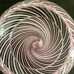 May include: A clear glass bowl featuring a swirling pattern of thin white and light pink stripes that radiate from the center toward the rim. The glass has a smooth, rounded interior surface and a circular shape, creating a vortex-like visual effect.