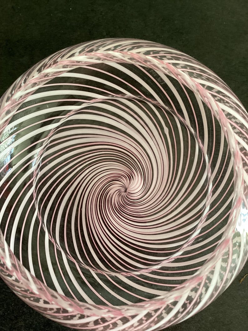 May include: A clear glass bowl featuring a swirling pattern of thin white and light pink stripes that radiate from the center toward the rim. The glass has a smooth, rounded interior surface and a circular shape, creating a vortex-like visual effect.