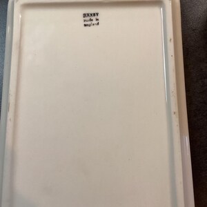May include: A rectangular white ceramic tray with a smooth, glossy finish. The underside features a black stamp with the text RAXBY and made in england. The tray has raised edges and a clean, simple design suitable for various household uses.