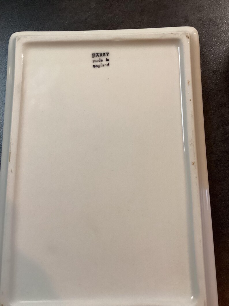 May include: A rectangular white ceramic tray with a smooth, glossy finish. The underside features a black stamp with the text RAXBY and made in england. The tray has raised edges and a clean, simple design suitable for various household uses.
