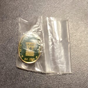 May include: A gold-coloured pin with a green centre, featuring the letters "KF" and a crown design. The pin is in a clear plastic bag. The text "KEVIN" and "Est. 1887" are visible on the pin.