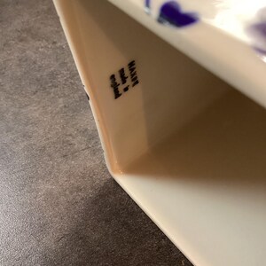 May include: A white ceramic vessel featuring a blue floral pattern on its exterior. The interior wall displays a black stamp with the text "MAXWELL made in england". The object has a smooth, glossy finish and a rectangular opening with rounded corners.