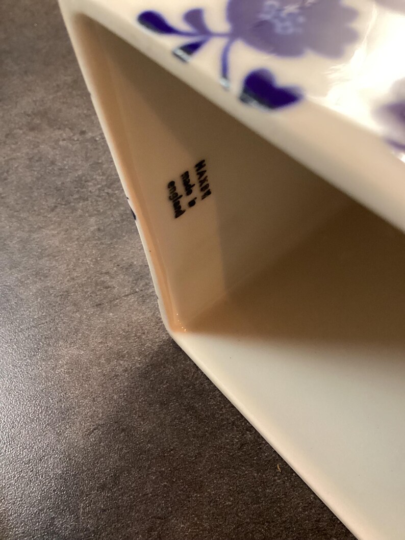 May include: A white ceramic vessel featuring a blue floral pattern on its exterior. The interior wall displays a black stamp with the text "MAXWELL made in england". The object has a smooth, glossy finish and a rectangular opening with rounded corners.