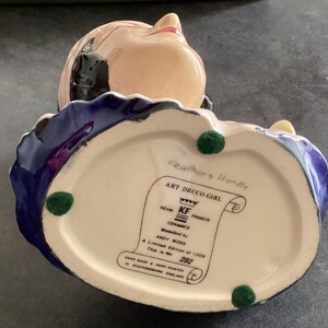 May include: A ceramic Art Deco Girl figurine with a cream base, blue and purple accents, and a peach face. The base has green felt feet and a label that reads "Art Decco Girl" and "Hand Made & Hand Painted in Staffordshire England."