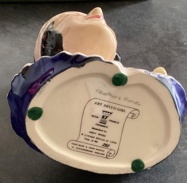 May include: A ceramic Art Deco Girl figurine with a cream base, blue and purple accents, and a peach face. The base has green felt feet and a label that reads "Art Decco Girl" and "Hand Made & Hand Painted in Staffordshire England."