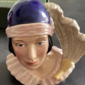 May include: A ceramic figurine of a woman's head, with a blue cap, pink headband, and a ruffled collar. The face has delicate features with painted eyes and lips, and a fan is held behind the head.