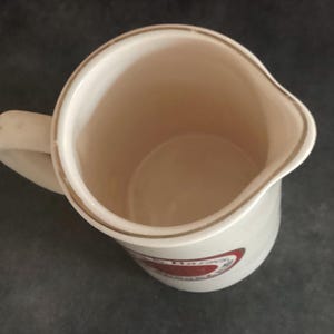 Op de afbeelding: Cr&egrave;mekleurige keramische kan met een handvat en een tuit. De kan heeft een goudkleurige rand rond de rand en een rood-wit ontwerp aan de zijkant. De binnenkant van de kan is lichtbeige.