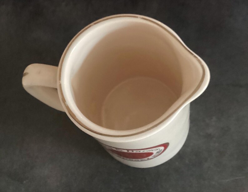 Op de afbeelding: Cr&egrave;mekleurige keramische kan met een handvat en een tuit. De kan heeft een goudkleurige rand rond de rand en een rood-wit ontwerp aan de zijkant. De binnenkant van de kan is lichtbeige.