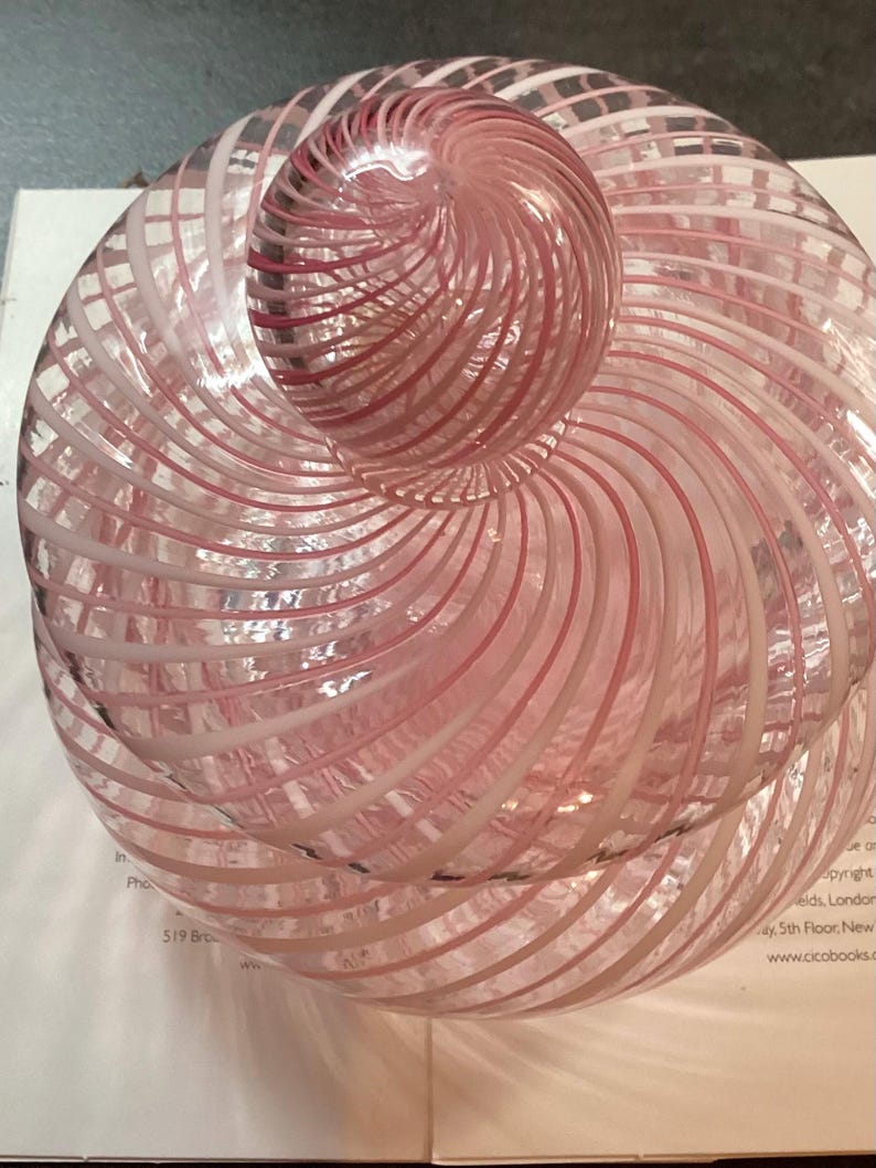 May include: A clear glass jar with a lid, featuring a swirling pattern of opaque white and translucent pink stripes. The jar has a rounded body and a matching spherical knob on top, with the glass design creating a continuous spiral effect.