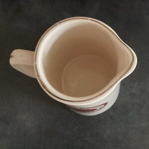 Op de afbeelding: Cr&egrave;mekleurige keramische kan met een handvat en een schenktuit. De rand is afgewerkt met een gouden band. Een rood-wit embleem is zichtbaar op de zijkant. De binnenkant is lichtbeige.