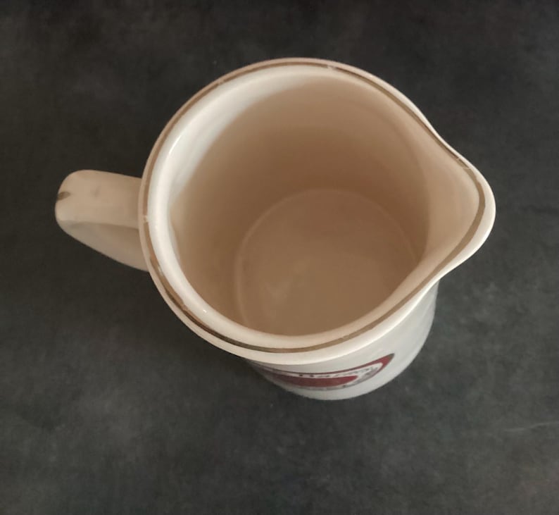 Op de afbeelding: Cr&egrave;mekleurige keramische kan met een handvat en een schenktuit. De rand is afgewerkt met een gouden band. Een rood-wit embleem is zichtbaar op de zijkant. De binnenkant is lichtbeige.