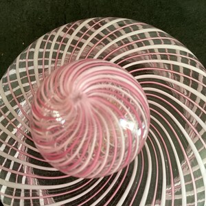 May include: A clear glass paperweight featuring a spherical top and a flat circular base, both decorated with an intricate pattern of swirling white and pink glass ribbons that radiate outward from the center in a continuous spiral design.