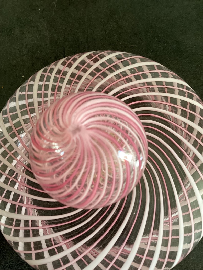 May include: A clear glass paperweight featuring a spherical top and a flat circular base, both decorated with an intricate pattern of swirling white and pink glass ribbons that radiate outward from the center in a continuous spiral design.