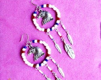 May include: A pair of handcrafted earrings featuring a silver eagle head pendant within a beaded hoop. The hoops are adorned with white, red, and blue beads, with dangling silver feathers. The earrings are set against a pink background.