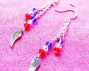 May include: Silver-toned dangle earrings with a patriotic theme. Each earring features a chain with red, white, and blue crystal beads and a small, detailed wing charm. The earrings are displayed on a pink surface.