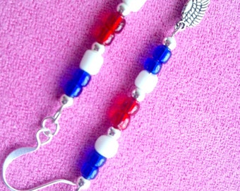 May include: A pair of dangle earrings with a patriotic theme. Each earring features a series of red, white, and blue beads, with a silver-colored wing charm at the bottom. The earrings are set against a pink background.