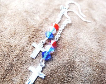 May include: Silver-toned earrings featuring two crosses and a chain with red and blue crystal beads. The earrings have a patriotic color scheme and a religious theme. The hooks are silver and the background is a brown, textured surface.