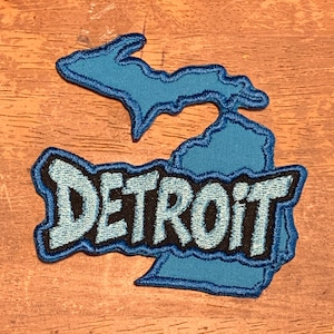 May include: A blue embroidered patch in the shape of the state of Michigan, with the word "DETROIT" in a stylized font. The patch has a black border and is set against a wooden background.