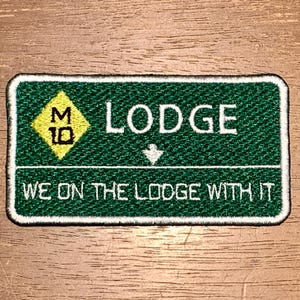 May include: A rectangular embroidered patch with a dark green background and a white border. It features a yellow diamond with the black text M 10, the white text LODGE, a small white downward arrow, and the white text WE ON THE LODGE WITH IT.