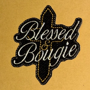 May include: A black embroidered patch in the shape of a cross featuring the white stitched text Blessed & Bougie. The patch has a gold-colored stitched border and gold-colored stitching outlining the cross shape against a solid black background.
