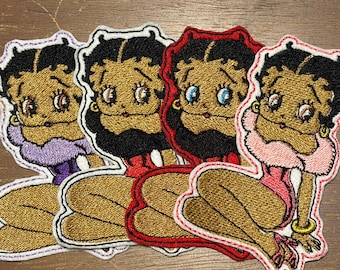 May include: Four embroidered patches featuring the cartoon character Betty Boop with black hair and gold skin. Each patch shows the character in a different colored outfit including purple, red, black, and pink, with matching colored stitched borders.