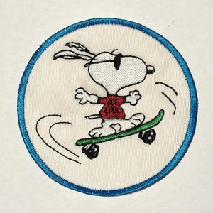May include: A circular embroidered patch featuring a white cartoon dog wearing black sunglasses and a red shirt with the text Joe Cool printed on it. The dog is riding a green skateboard, and the entire patch is bordered by a bright blue stitched edge.