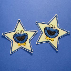 May include: Two embroidered star-shaped patches featuring a light yellow center with a blue border. Each patch displays a blue fuzzy character face with large white eyes, a wide black mouth, and a small yellow bow tie at the bottom.