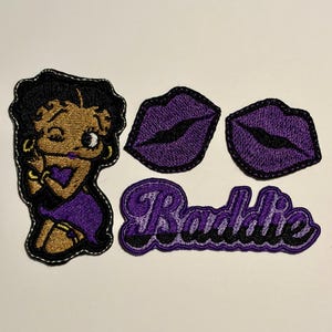 May include: A set of four embroidered patches featuring a cartoon character with black hair wearing a purple dress, two purple lips, and a purple script patch with the word Baddie in black lettering, all with black stitched borders.