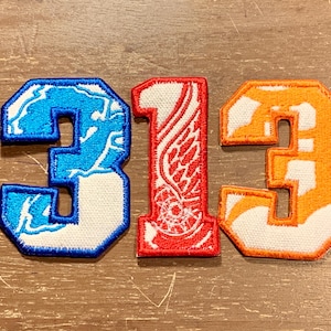May include: Three embroidered patches in the shape of the number 3 and 1. The number 3 is in blue with a white lightning pattern, and orange with a white outline. The number 1 is red with a white wing design.